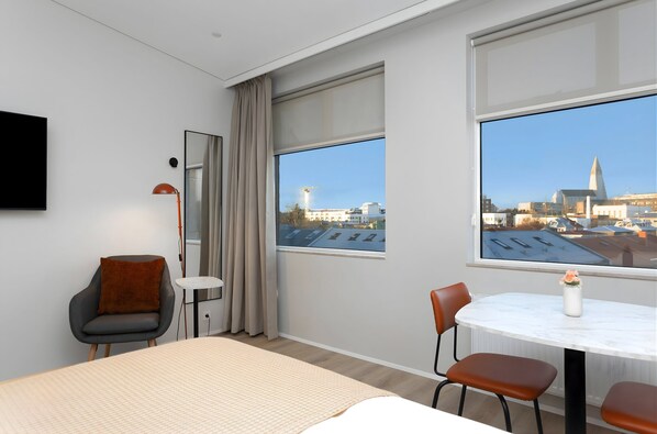Una habitación de hotel moderna con una cama, una mesa de comedor redonda con sillas, una mesita auxiliar pequeña, una silla con cojín, un televisor montado en la pared y grandes ventanales con vistas a la ciudad.