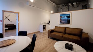 Un salon moderne avec un canapé en cuir marron, une table à manger blanche avec un plateau en bois et une petite table blanche sur laquelle se trouve un objet noir. Il y a un mur blanc orné d’un tableau encadré et un meuble blanc avec un évier.