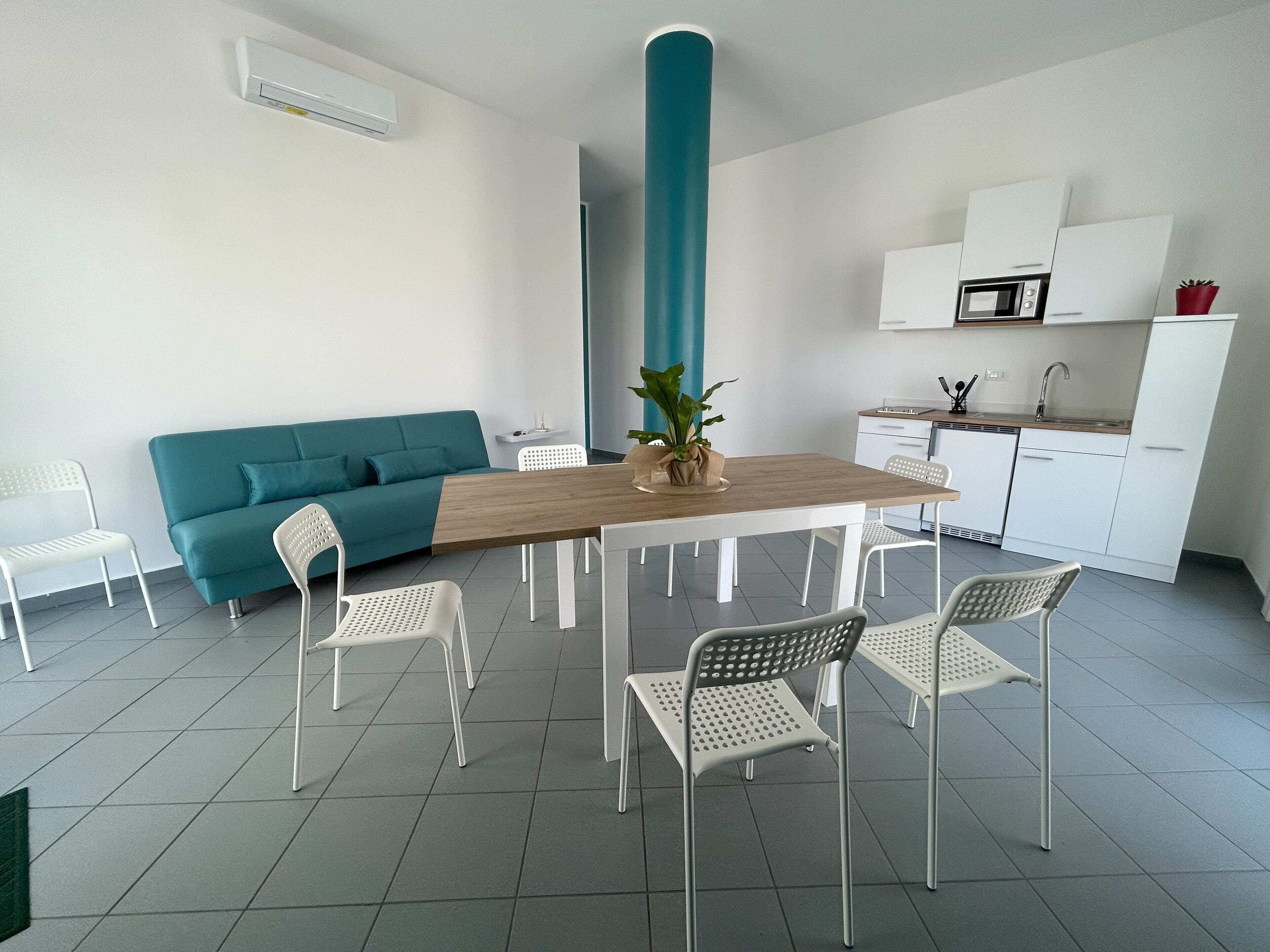Una cocina moderna con armarios blancos, un pilar verde azulado y una mesa de comedor de madera rodeada de sillas blancas.