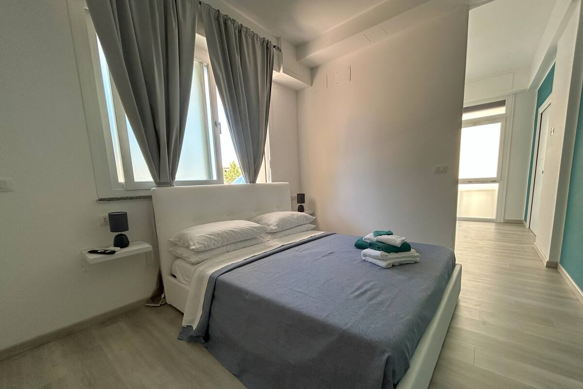 Een moderne slaapkamer met een bed, gordijnen, een nachtkastje en uitzicht op een gang.