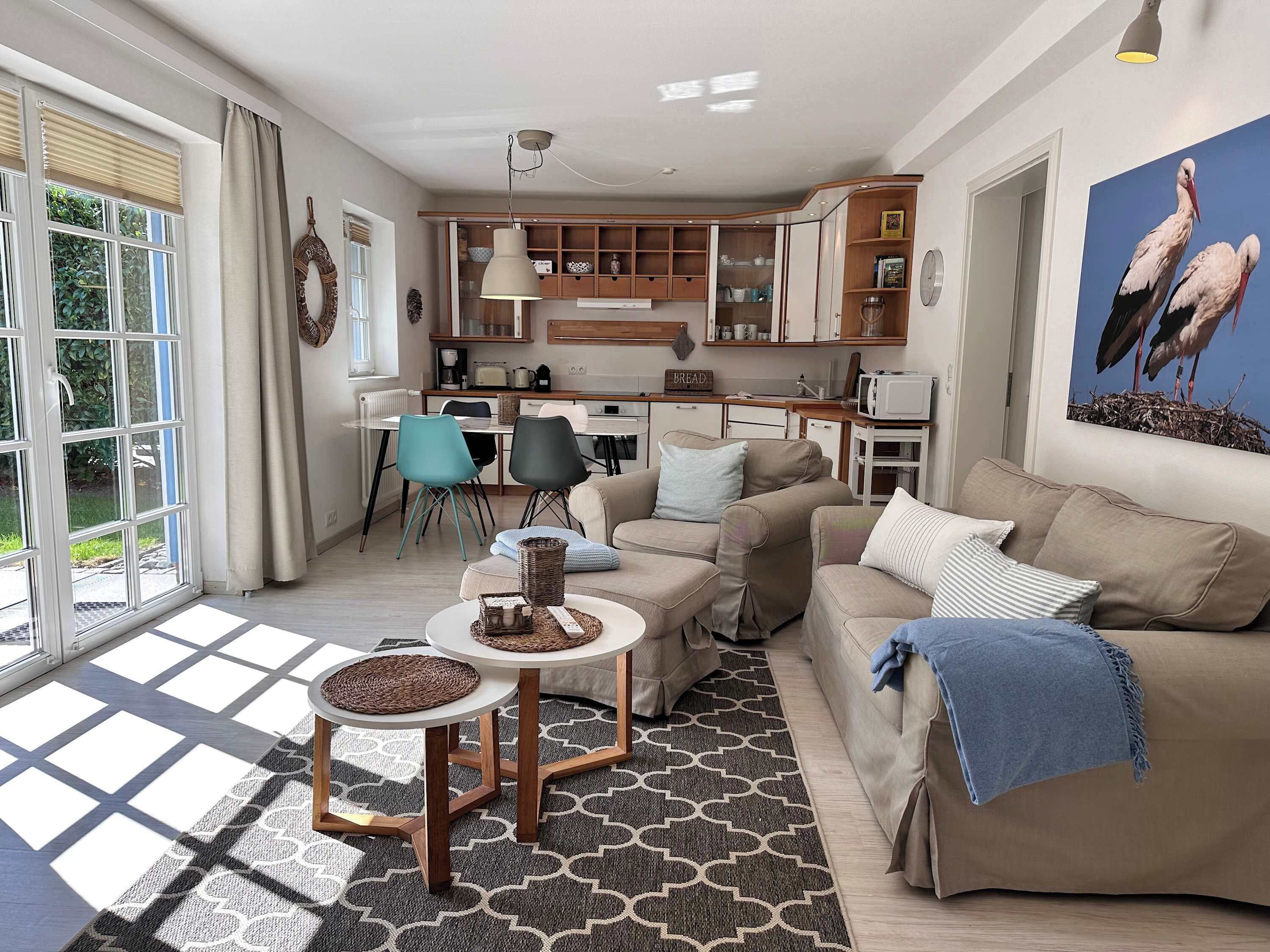 A modern living room with a sofa, coffee table, and chairs. There is a kitchen area with cabinets and a microwave. A large window with blinds is visible, and a painting of storks is on the wall.