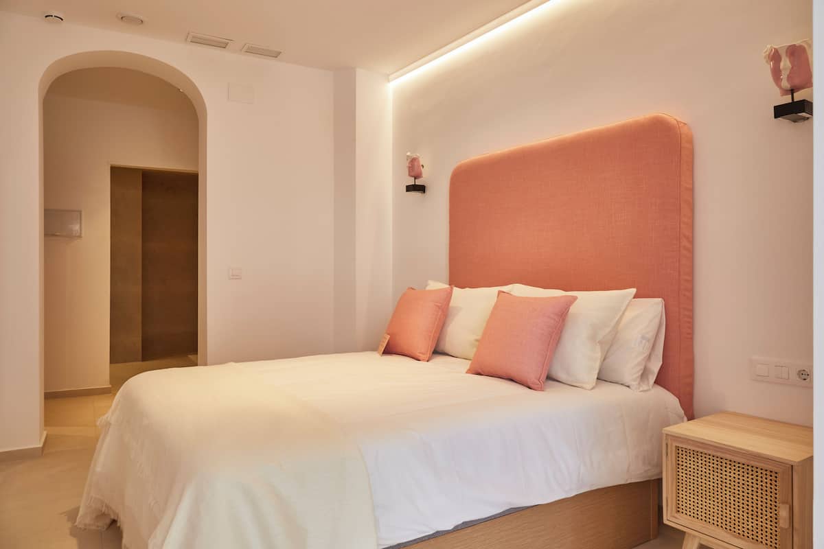 A bedroom with a bed, a nightstand, a wall-mounted shelf, and a door leading to another room.