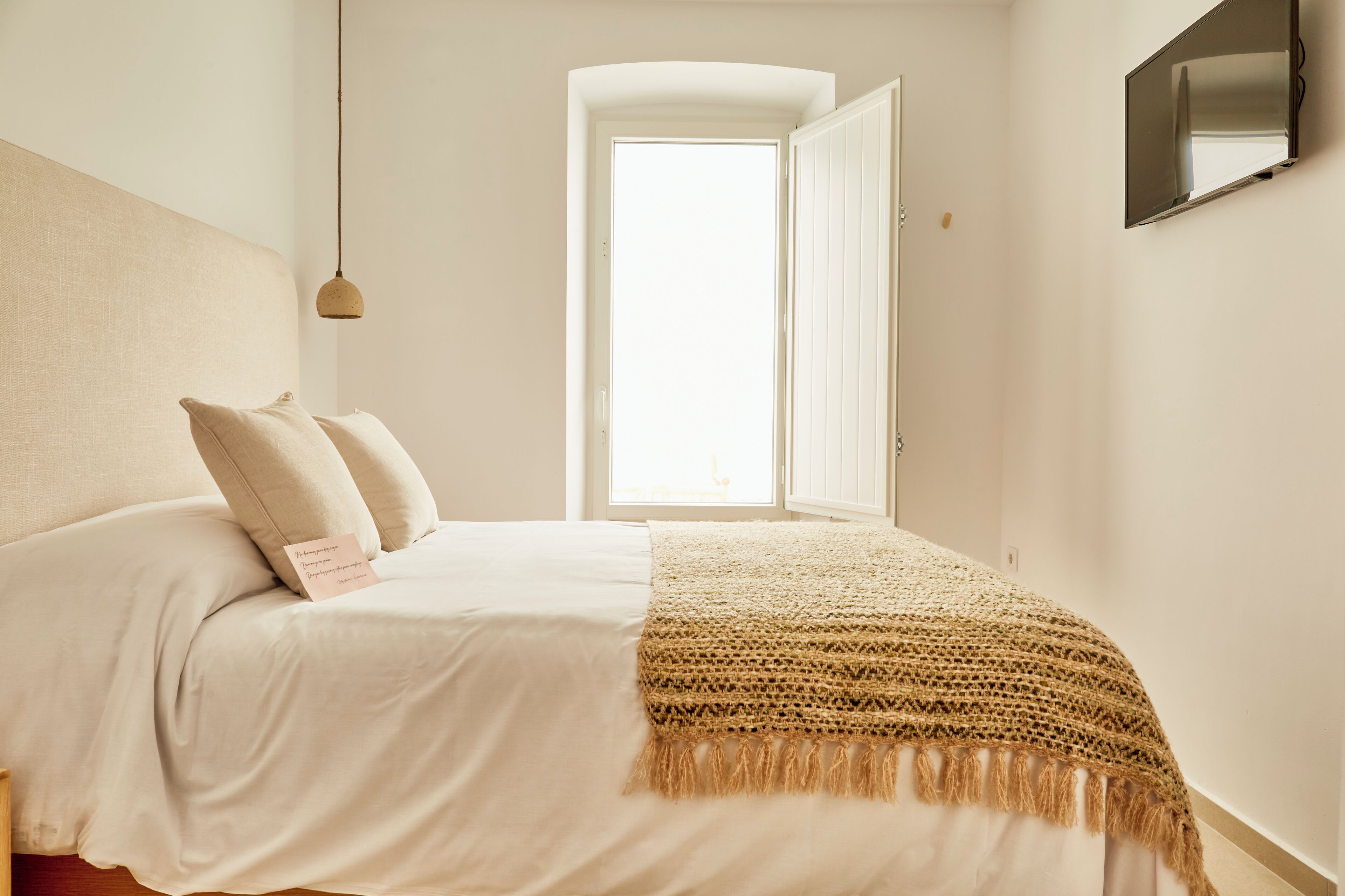 Un dormitorio con cama, almohadas, un plaid de punto, una lámpara colgante y una ventana con cortinas.