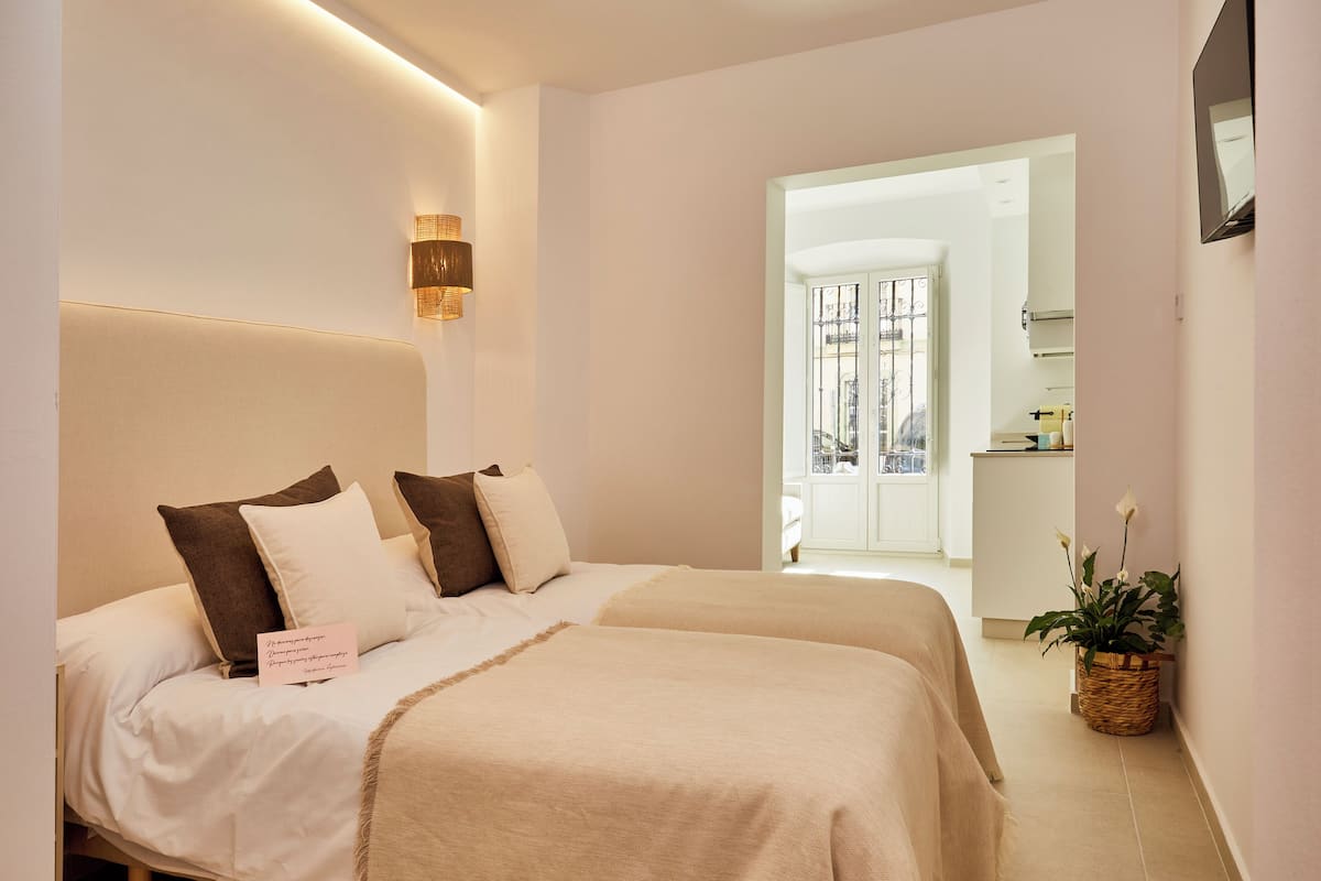 A bedroom with two beds, a bedside lamp, a bedside plant, and a view of the kitchen and balcony.