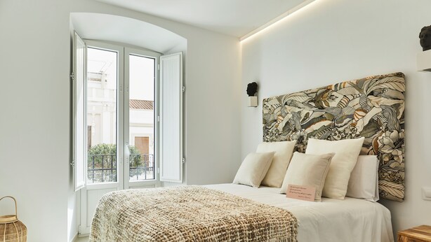 Une chambre avec un grand lit, une couverture en tissu tissé et une vue sur l’extérieur par une porte ouverte.