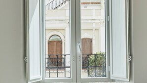 Une pièce dotée d’un grand tapis à motifs, d’une porte-fenêtre coulissante donnant sur un balcon extérieur avec une rambarde en métal, et offrant une vue sur un bâtiment avec une porte en bois et un balcon.