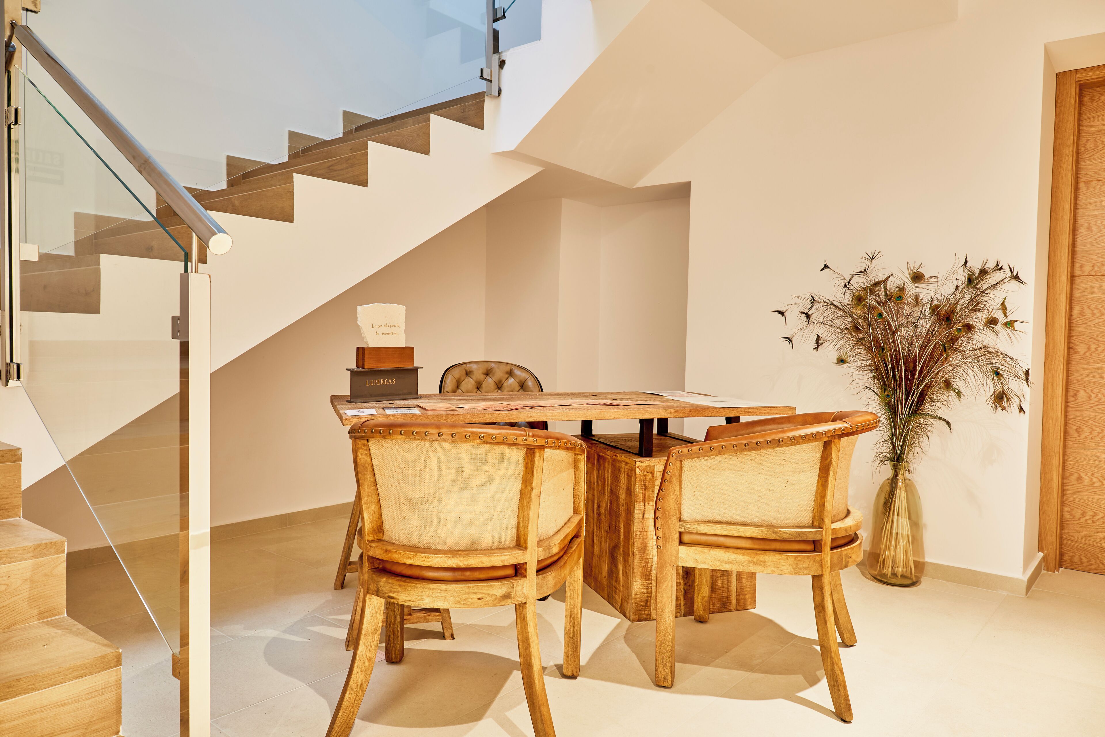A modern dining area with a wooden table and chairs, a vase with dried plants, and a staircase in the background.