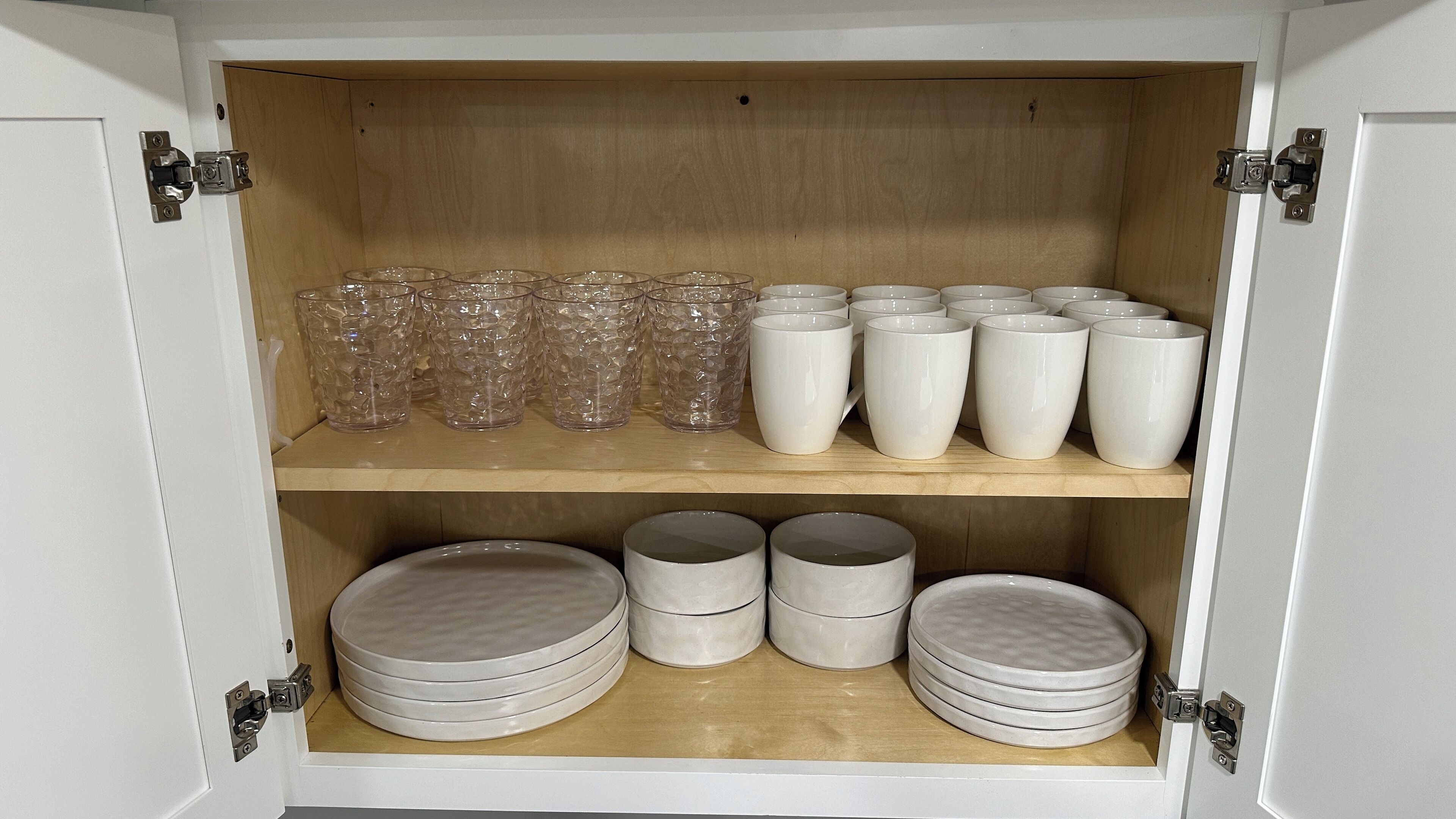 A cabinet with shelves containing clear glass tumblers, white mugs, and stacked plates.