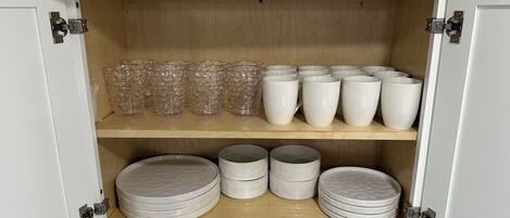 A cabinet with shelves containing clear glass tumblers, white mugs, and stacked plates.