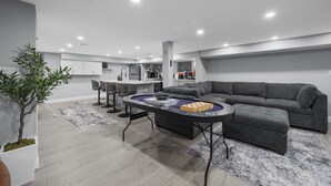 Un saló modern amb un sofà gris, una taula de centre amb un joc de taula, una zona de menjador amb cadires, i una cuina al fons.