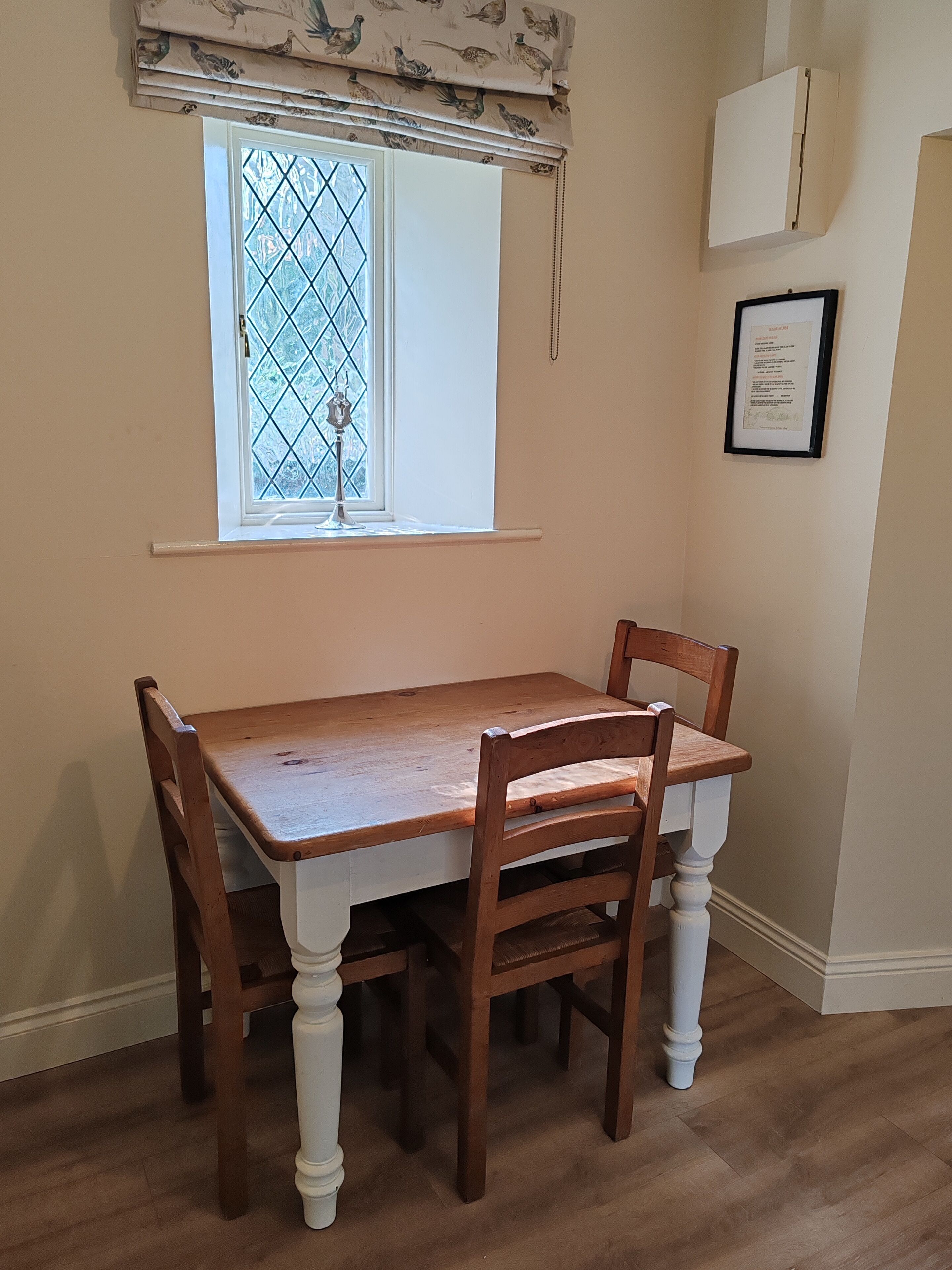 A dining area with a wooden table and chairs, a window with a decorative valance, and a framed certificate on the wall.