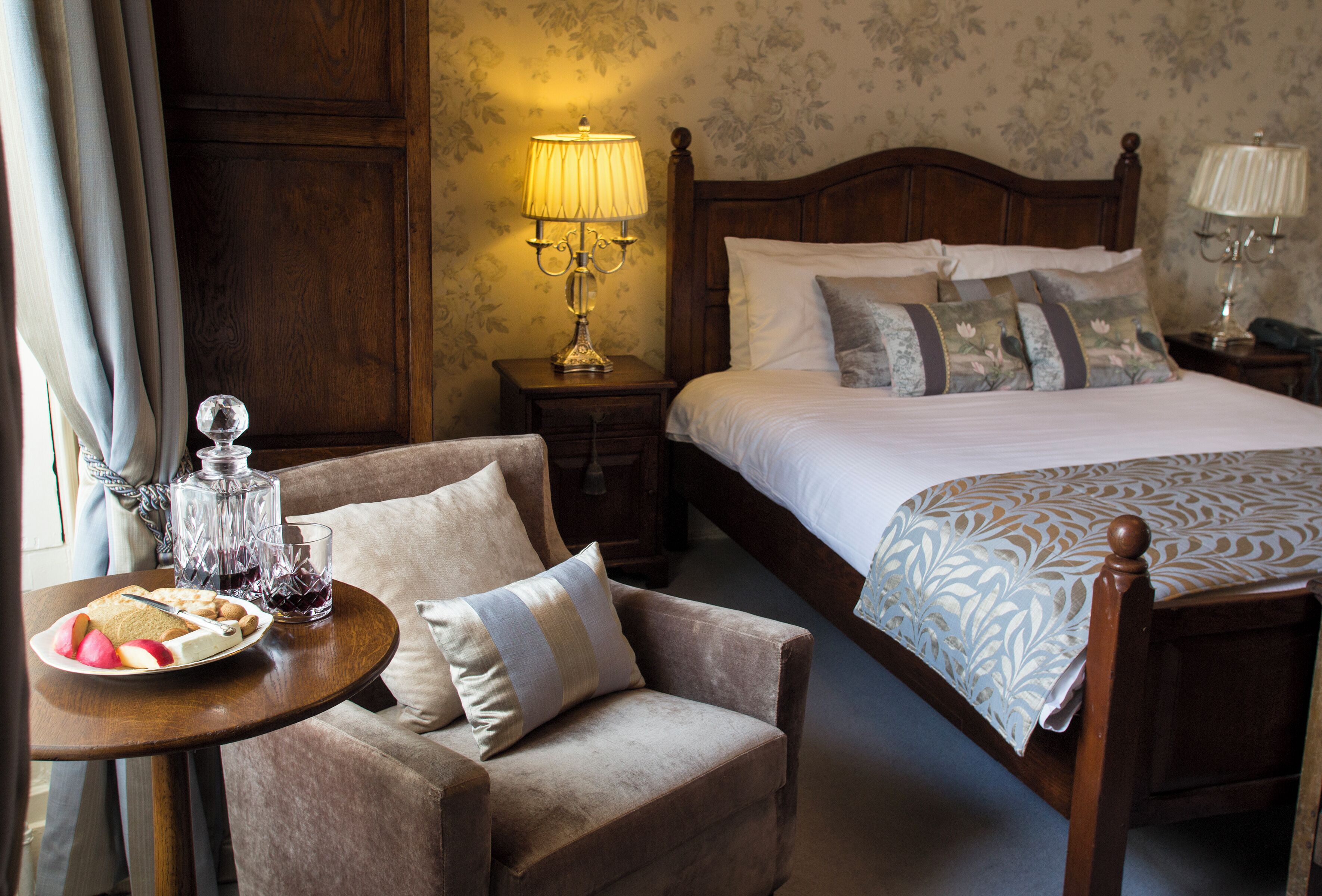 A hotel room with a wooden bed, a bedside table with a lamp, a chair, and a table with food and drinks.