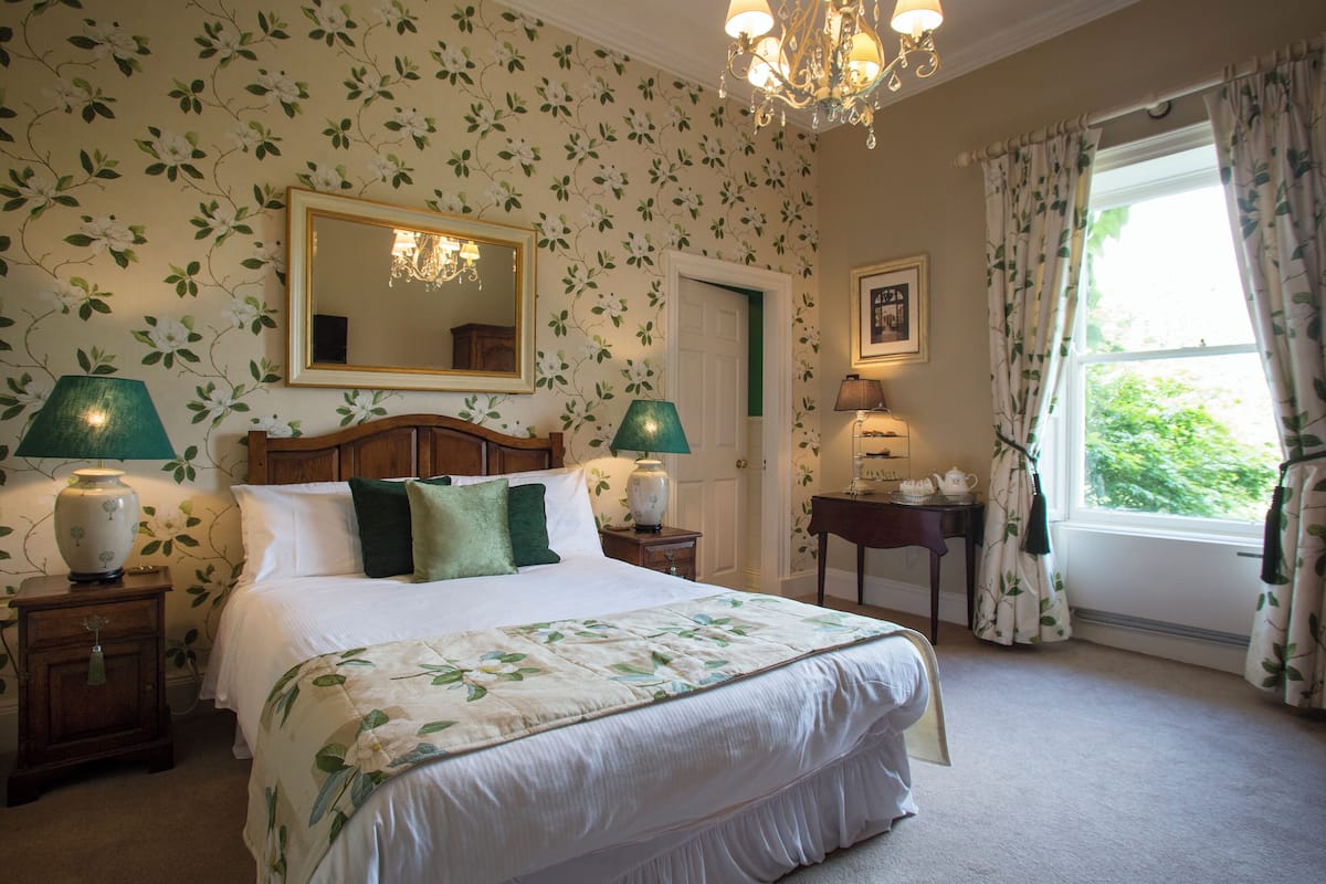 A bedroom with a large bed, floral wallpaper, a chandelier, and a window with curtains.