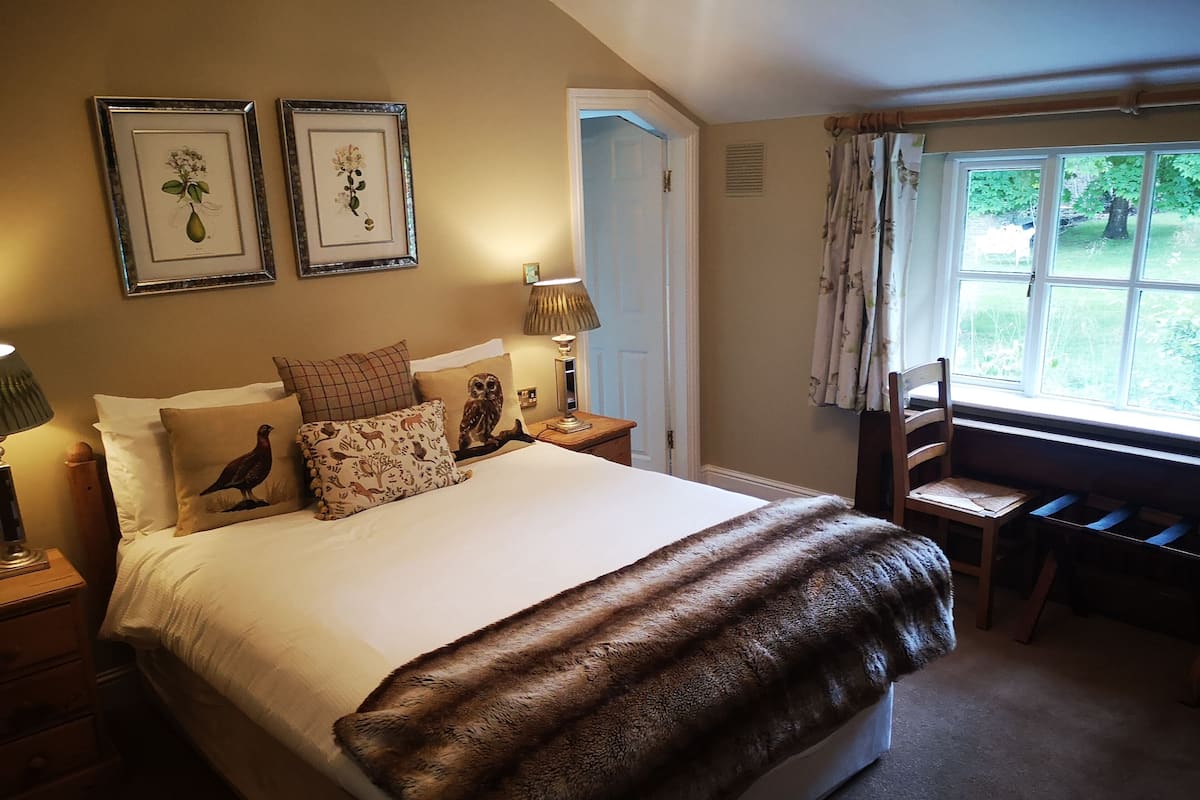 A neatly arranged bedroom with a large bed, two framed floral pictures, a wooden chair, and a window with a view of greenery.