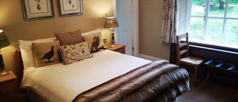 Un dormitorio ordenado con una cama grande, dos cuadros enmarcados con motivos florales, una silla de madera y una ventana con vistas a zonas verdes.