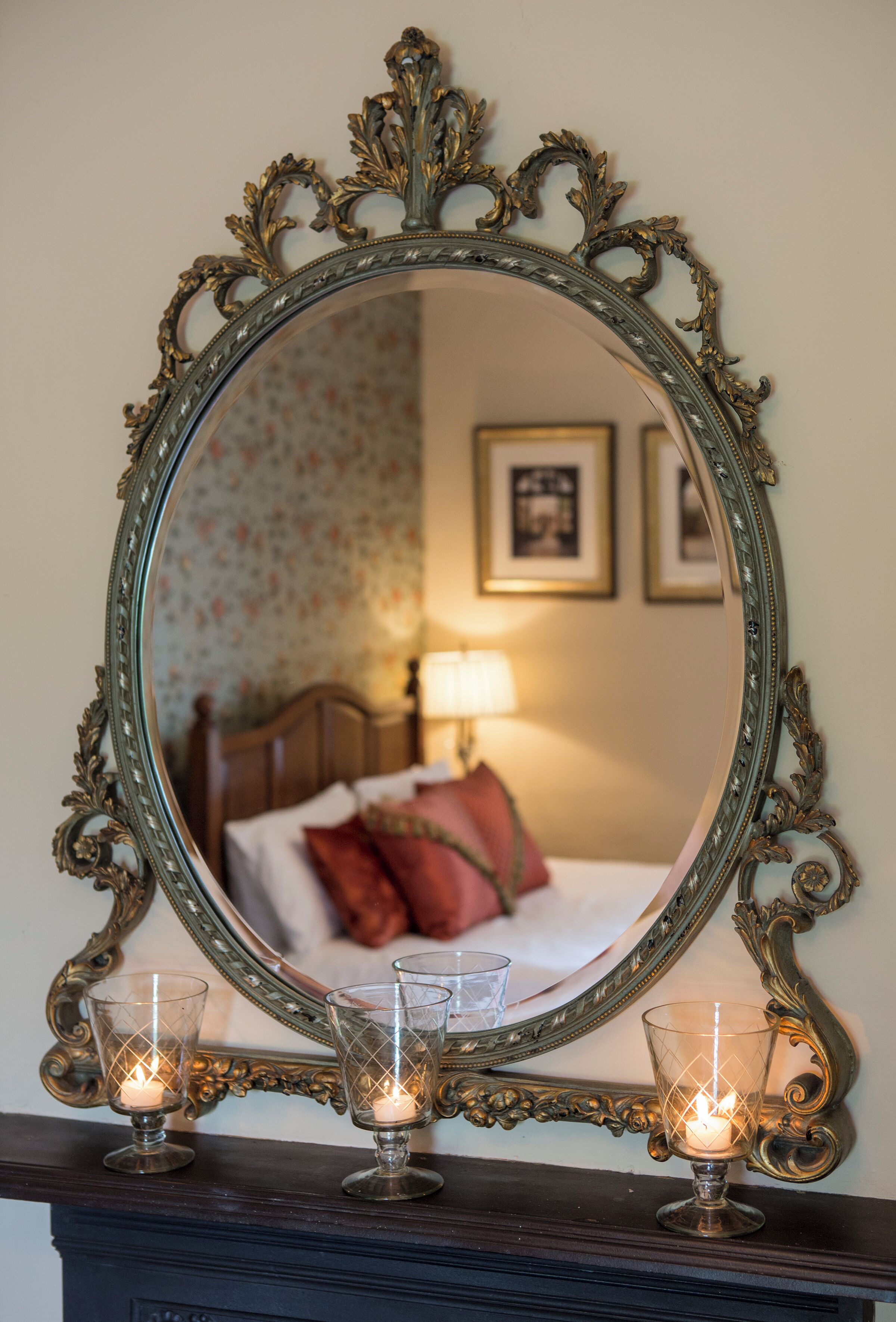 A mirror with an ornate frame, two candle holders with candles, and a bed with pillows in the background.