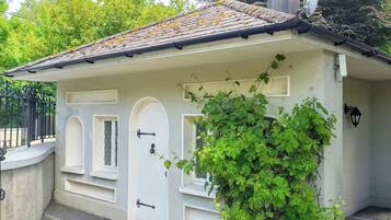 A small, single-story building with a white facade, a tiled roof, and a small garden area with green foliage.