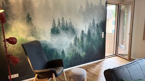 A room with a forest mural, a red floor lamp, a grey armchair, a small ottoman, and a black leather sofa with a green cushion. - Family-friendly vacation apartment - four-legged friends also welcome! (Binzen)