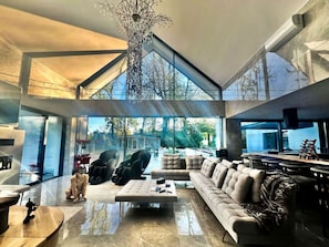 A spacious living area with a high ceiling, large windows, and a modern chandelier.