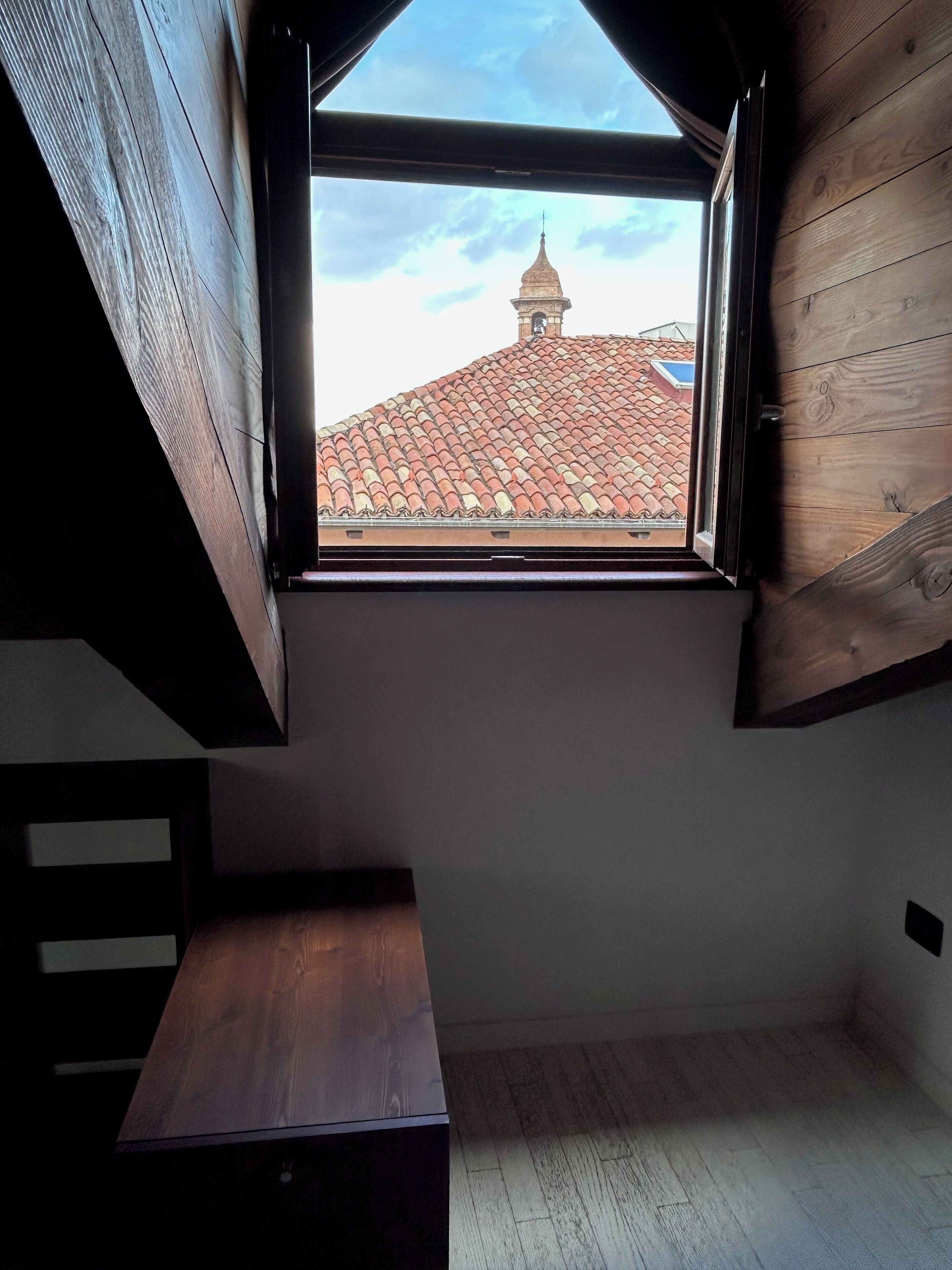 A room with a sloped ceiling and a window overlooking a tiled roof with a chimney.