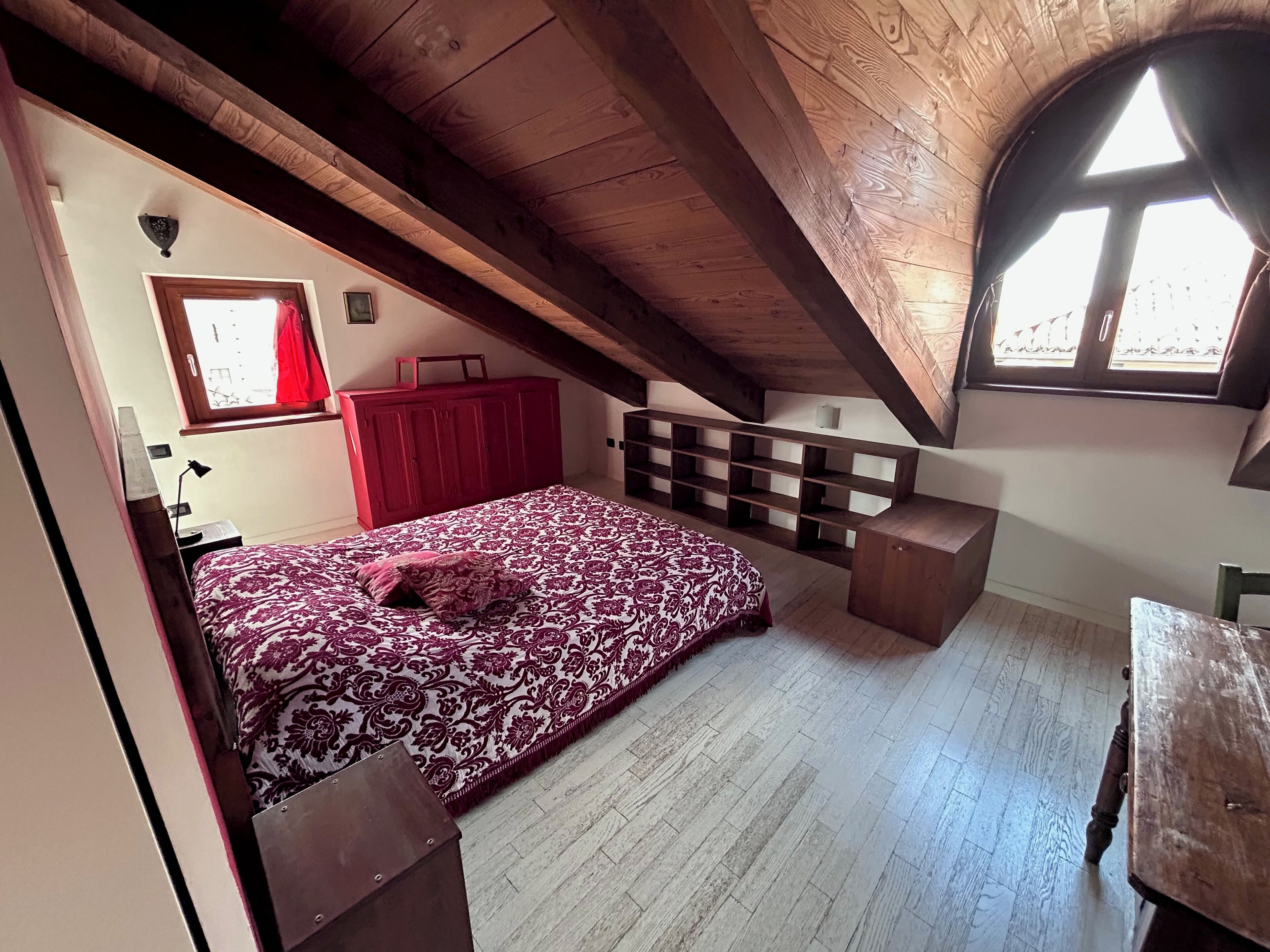 A bedroom with a sloped ceiling, a bed with a patterned bedspread, a red wardrobe, and a window with a view.