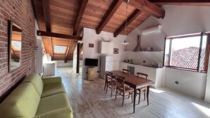 A spacious loft with exposed wooden beams, a dining area with a wooden table and chairs, a green sofa, a kitchen with white cabinets, and a large window with a view of a tiled roof.