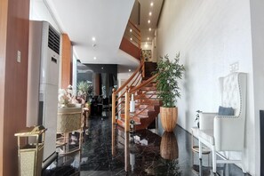 Un moderno vestíbulo de hotel con suelo de mármol, una escalera de madera, una gran planta en maceta y un sillón blanco tapizado.