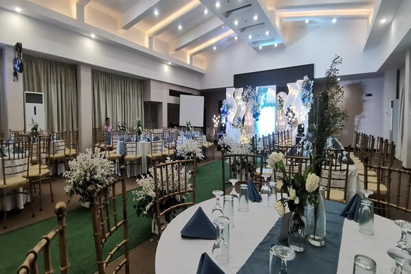 Un salón de banquetes con mesas redondas preparadas para eventos, con centros de mesa florales blancos y servilletas azules.