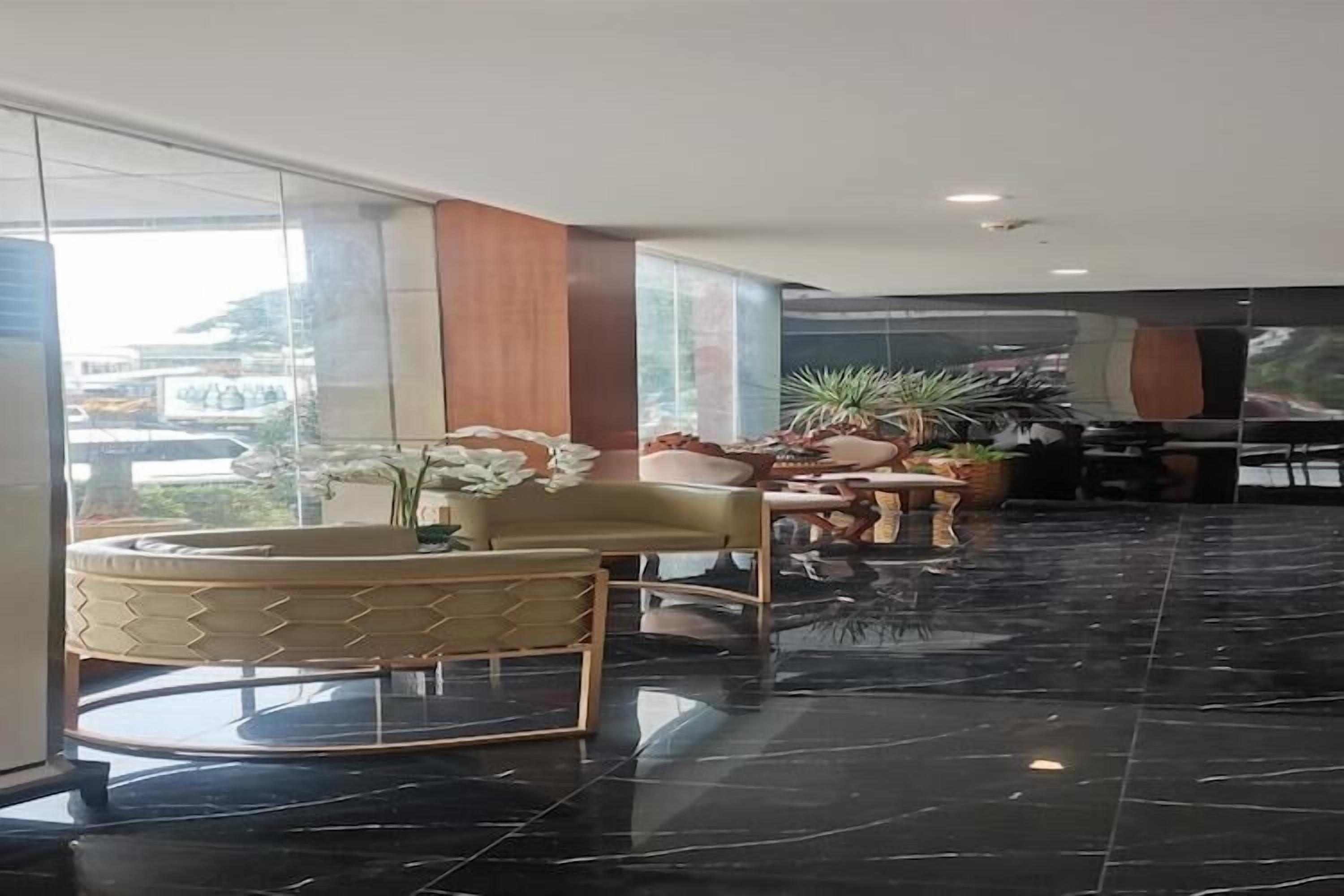Een moderne lobby met een vloer van zwart marmer, een ronde houten tafel en een bank met een gestreept kussen. Er staan potplanten en een groot raam dat uitzicht biedt op buiten.