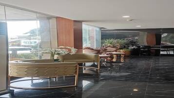 Een moderne lobby met een vloer van zwart marmer, een ronde houten tafel en een bank met een gestreept kussen. Er staan potplanten en een groot raam dat uitzicht biedt op buiten.