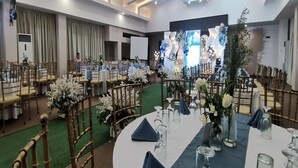 A banquet hall with round tables set for events, featuring floral centerpieces and a stage with a backdrop.