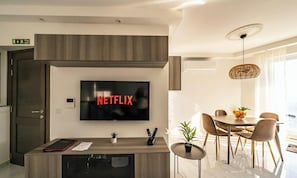 Un soggiorno moderno con una TV a schermo piatto fissata al muro che mostra 'NETFLIX', un mobiletto in legno, un tavolino con una pianta, e un'area pranzo con sedie e un tavolo.