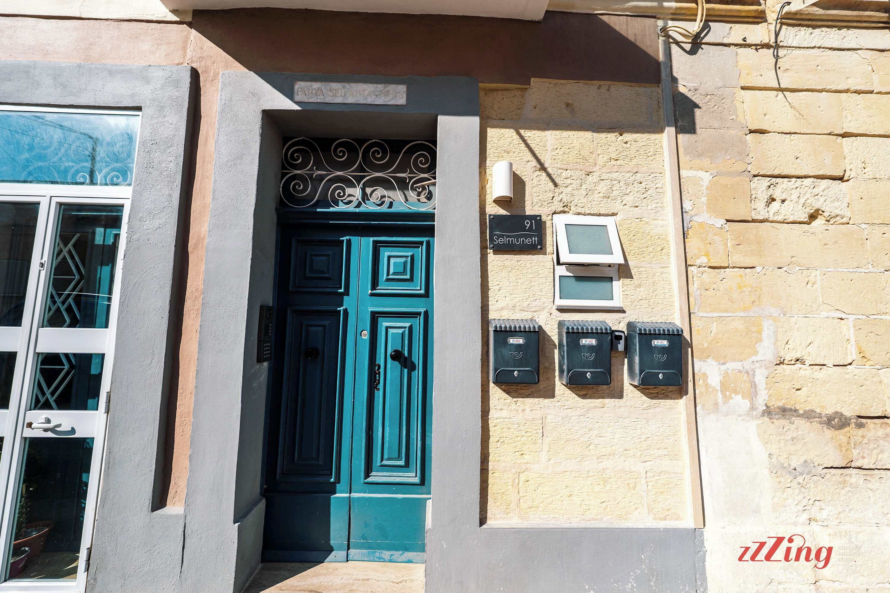 Entrada de un edificio con una puerta verde azulada, un buzón y un letrero que dice "91 Selmunett".