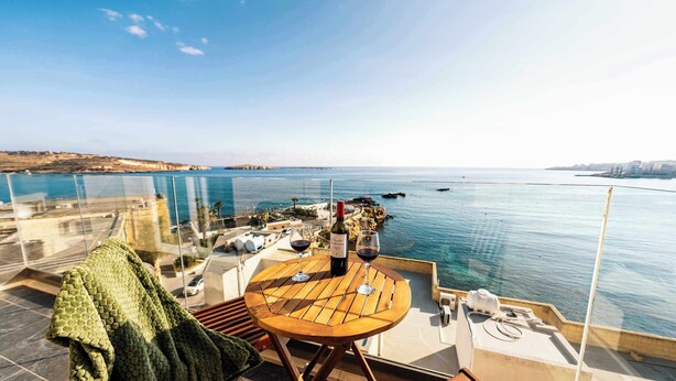 A balcony with a table, two wine glasses, and a bottle of wine. The view includes a body of water and distant land. - Seaside Serenity at 2BR home in San Pawl il-Baħar (San Pawl il-Baħar)