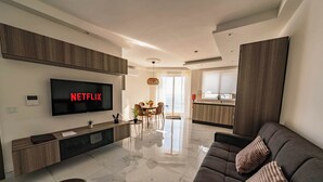 A modern living room with a flat-screen TV displaying 'NETFLIX', a dining area with a table and chairs, and a kitchen with a window view. - Seaside Serenity at 2BR home in San Pawl il-Baħar (San Pawl il-Baħar)