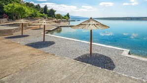 Uma área à beira da piscina com água azul cristalina, rodeada por chão de pedrinhas e protegida por guarda-sóis de palha.