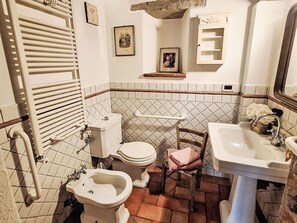 Een badkamer met toilet, bidet, wastafel en spiegel. Er hangen foto’s aan de muur en boven de wastafel is een plank.