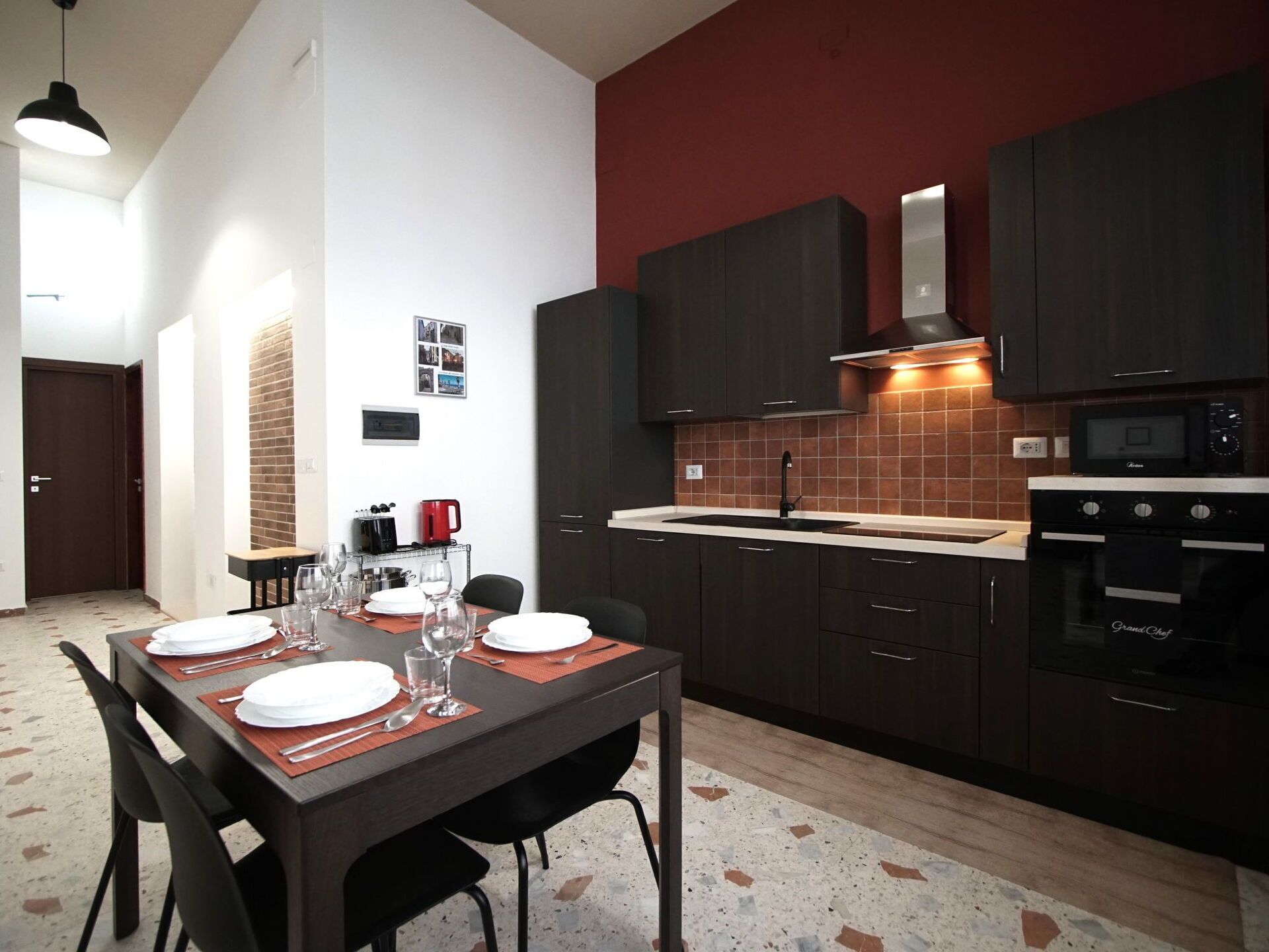 A modern kitchen with dark cabinets, a red accent wall, and a dining area set for four with a table, chairs, and a coffee machine.