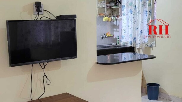 A room with a flat-screen TV mounted on the wall, a small wooden table, a sink with a mirror, and a blue trash bin. - 1BHK apartment in a Holiday Resort close to the Beach with a common pool (Calangute)