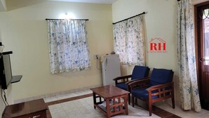 A living room with a wooden table, two blue chairs, a small cabinet, and a television. - 1BHK apartment in a Holiday Resort close to the Beach with a common pool (Calangute)