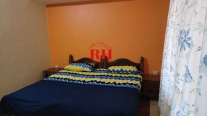 A bedroom with a bed, a nightstand, a wooden headboard, and a floral curtain. - 1BHK apartment in a Holiday Resort close to the Beach with a common pool (Calangute)