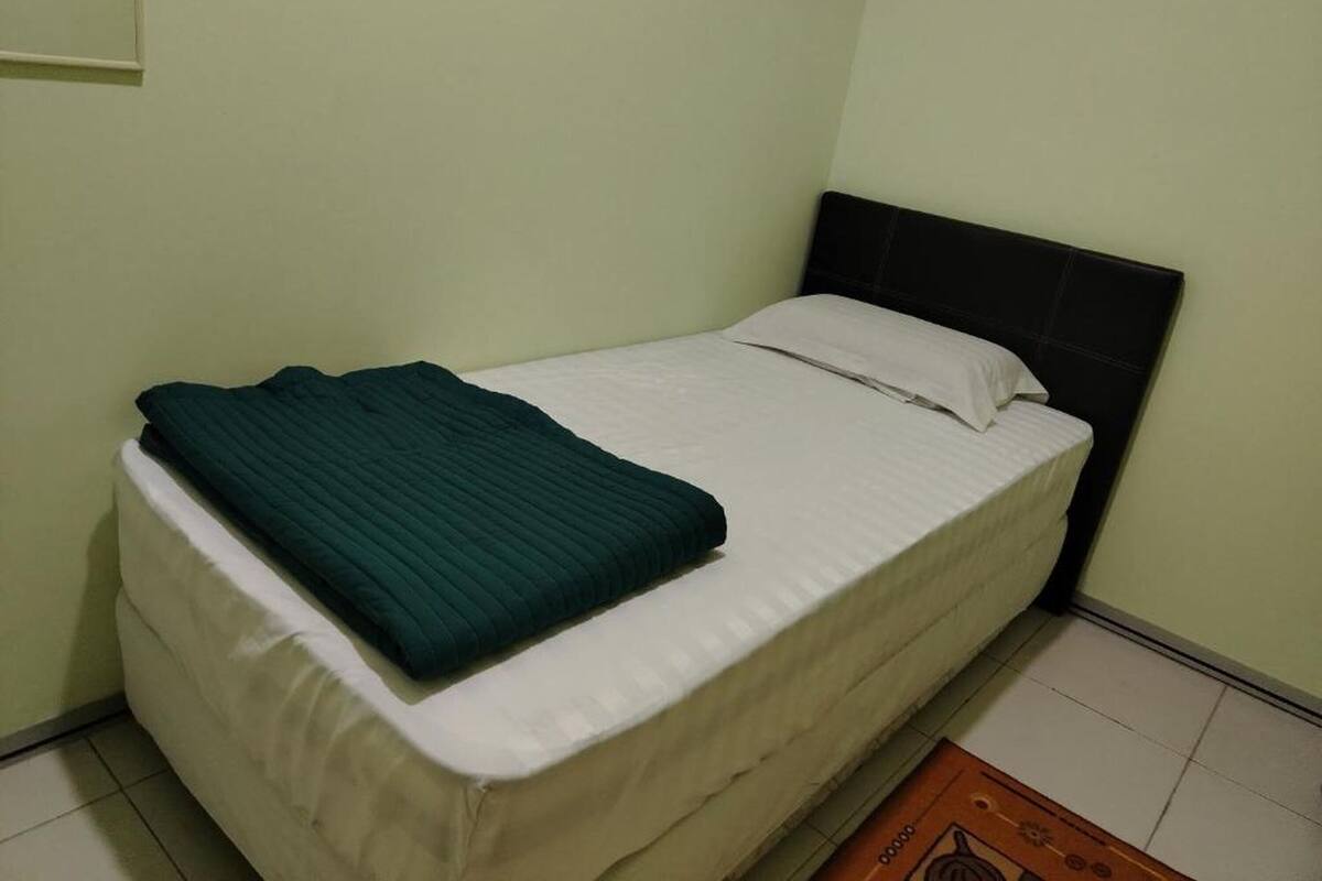 Un lit simple avec un matelas blanc et une couverture vert foncé, une tête de lit noire et un mur vert clair.