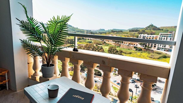 Un balcon agrémenté d’un pot de fleurs, une table avec un carnet et une tasse, et une vue sur un paysage urbain avec des bâtiments et des collines.