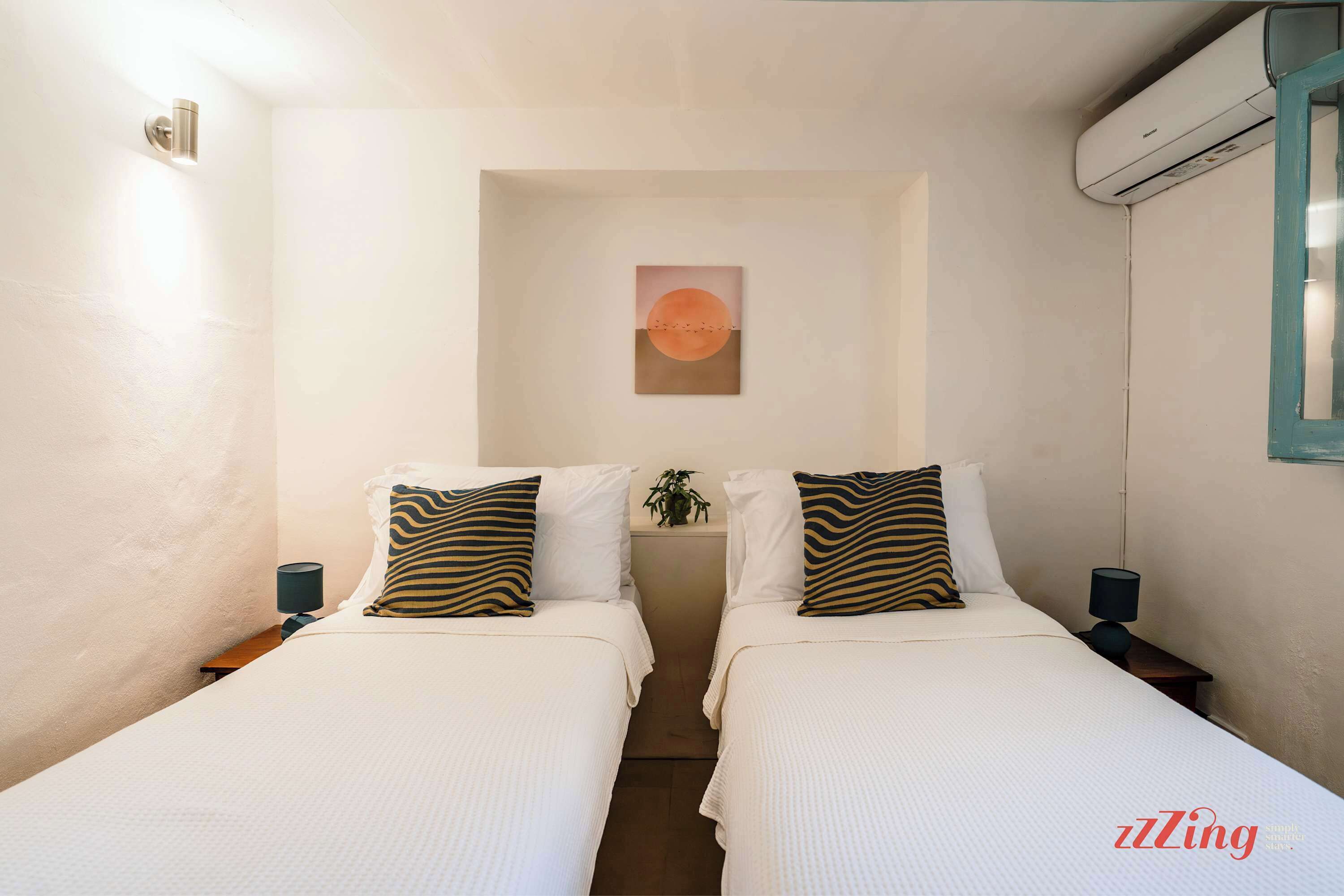 A hotel room with two beds, a wall-mounted air conditioner, a small plant, and a painting of a sun.