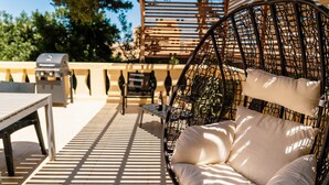 Un espacio exterior con asientos de mimbre, una silla acolchada y una mesa, una parrilla y una pérgola.