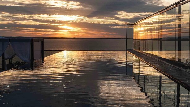 A waterfront deck with a glass railing overlooking the sea, a sunset in the background, and a clear sky. - Alula Hotel Encarnacion (Encarnación)