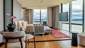 A modern hotel room with a large window offering a city view, a flat-screen TV, a dining area with a table and chairs, and a cozy seating area with cushions. - Alula Hotel Encarnacion (Encarnación)