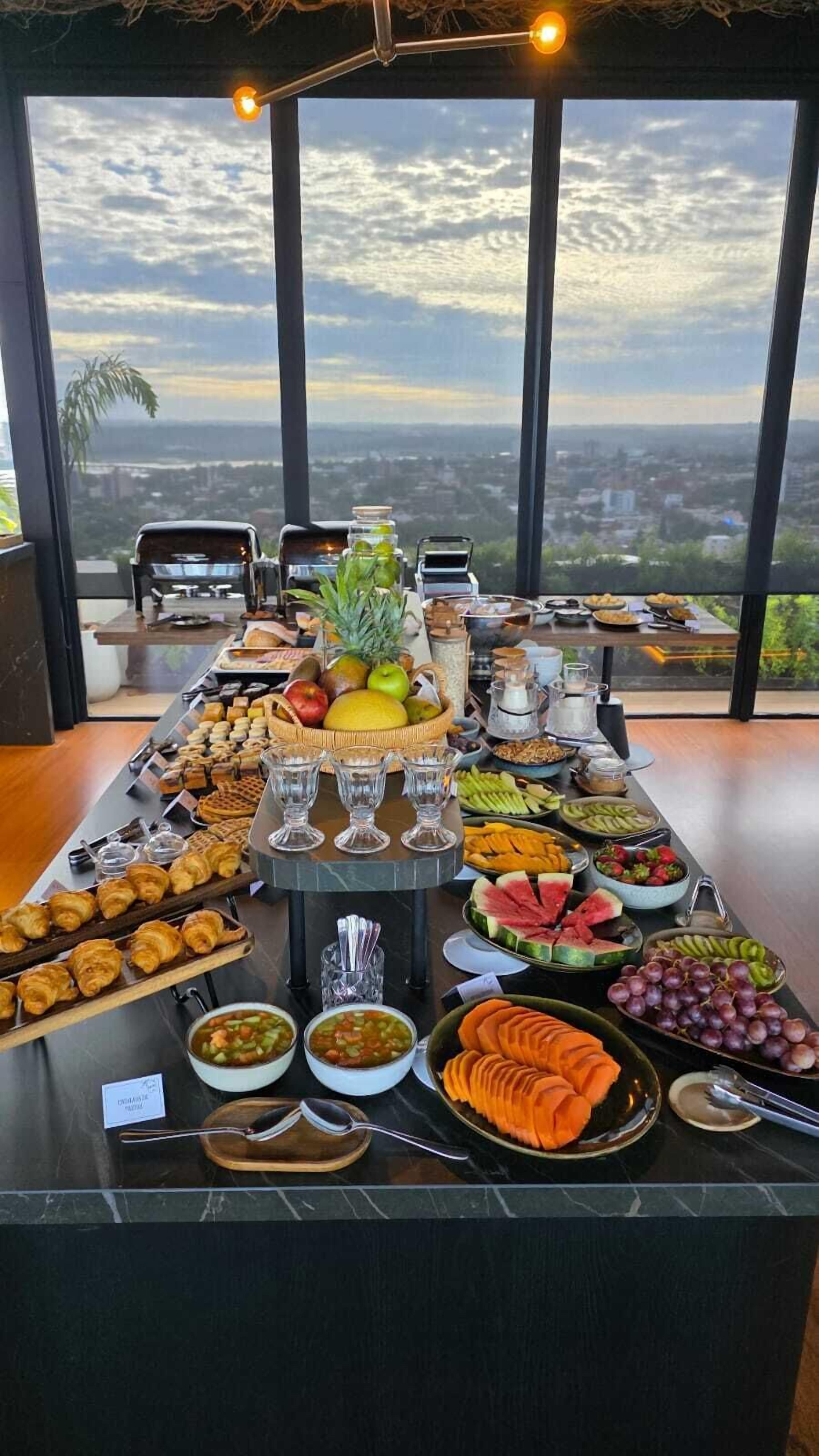A buffet spread with various fruits, pastries, and beverages on a table with a city view.