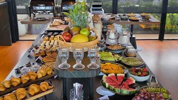 A buffet spread with various fruits, pastries, and beverages on a table with a city view.