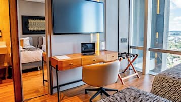 A modern bedroom with a wooden desk, a laptop, a TV, and a large window offering a city view.